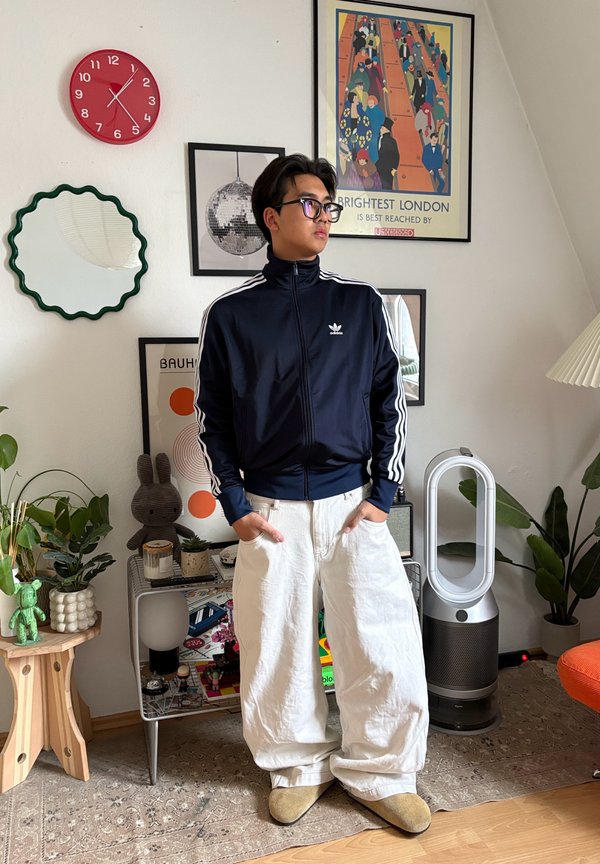 Junger Mann in schwarzer Adidas-Jacke und weißen Hosen steht in einem dekorierten Raum mit Pflanzen, einer roten Uhr, gerahmter Kunst und Haushaltsgeräten.