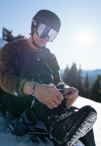 Personne portant un casque et des lunettes de ski attachant des chaussures de snowboard noires en s'asseyant sur la neige avec des arbres et le soleil en arrière-plan.