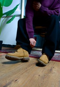 Braune Wildlederstiefel mit Gummisohlen, gelben Schnürsenkeln und verstärktem Zehenbereich. Person passt ihre Hose an, während sie auf einem Holzboden sitzt.