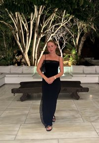 Vestido negro con detalles de encaje, diseño sencillo y tirantes ajustables; combinado con sandalias negras. Suelo de mármol y banco de madera en el fondo.
