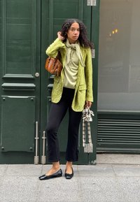 Vrouw met krullend haar in een groene blazer, lichtgroene top, zwarte broek, draagt een bruine handtas over de schouder en houdt een geruite parasol vast.