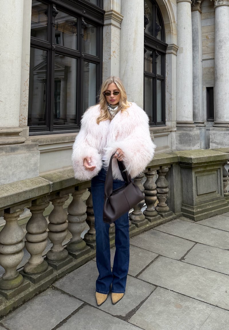 Γυναίκα με αφράτο ανοιχτό ροζ μπουφάν, μπλε τζιν, κίτρινα μυτερά παπούτσια και καφέ γυαλιά ηλίου, κρατώντας μια σκούρα καφέ τσάντα σε πέτρινο μπαλκόνι.