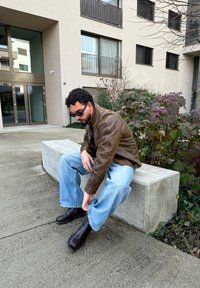 Veste en cuir marron sur une chemise en denim bleu, jeans larges bleu clair, et bottines marron. Cadre extérieur avec une architecture moderne.