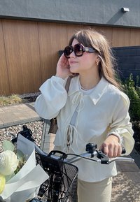 Vit, lätt blus med puffärmar och knytframsdesign; beige byxor. Svart cykel med en nätkorg som håller blommor.