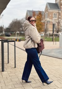 Beige Kapuzenjacke aus Kunstfell, dunkelblaue Jeans mit weitem Bein und schwarze spitze High Heels. Accessoires sind übergroße schwarze Sonnenbrillen und eine burgunderrote Tasche.