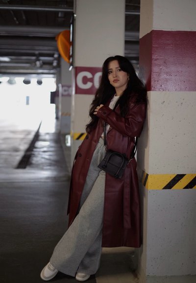 Abrigo de cuero burdeos, pantalones de pierna ancha grises, zapatillas blancas y bolso pequeño negro. Está de pie contra un pilar en un aparcamiento.