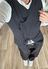 Persona con un top avvolgente nero senza maniche a strati sopra una maglietta bianca a maniche lunghe, pantaloni neri e sneakers bianche, in piedi su un pavimento di legno.