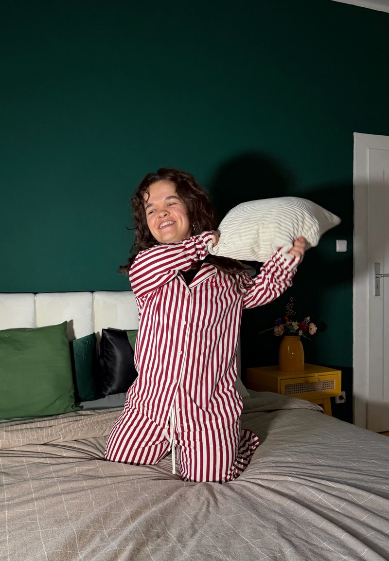 Jeune personne en pyjama rayé rouge et blanc à genoux sur le lit, tenant un oreiller prêt à être lancé, avec un mur vert foncé et une table de chevet en arrière-plan.