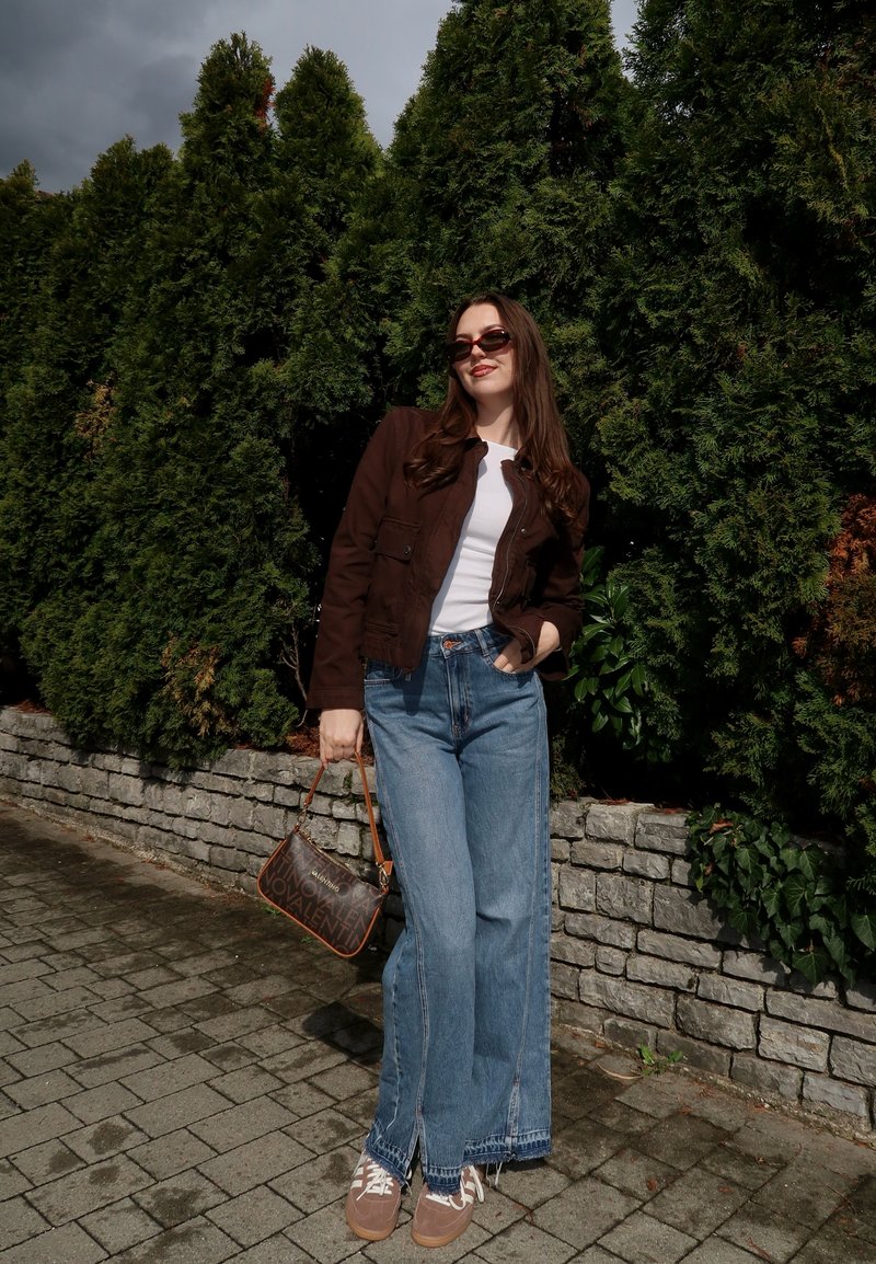 Woman wearing sunglasses, brown jacket, white shirt, wide-leg jeans, and brown sneakers holds a small handbag standing by a stone wall and greenery.