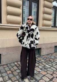 Femme debout sur une rue pavée portant un manteau en fausse fourrure à taches noires et blanches, un pantalon large noir, des lunettes de soleil, avec un petit sac à main marron.
