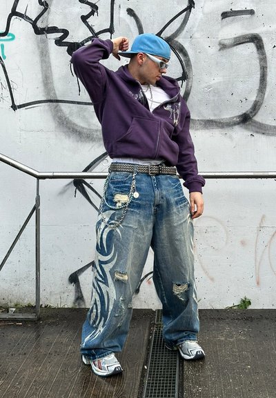 Sudadera con capucha morada, jeans desgastados de color azul con un gran diseño gráfico, cinturón de cadena y zapatillas grises con detalles negros.