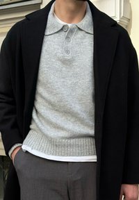 Vīrietis, tērpies pelēkā adītā džemperī ar apkakli un pogām, baltā krekliņā, melnā mētelī un pelēkās biksēs, stāv ar vienu roku kabatā.