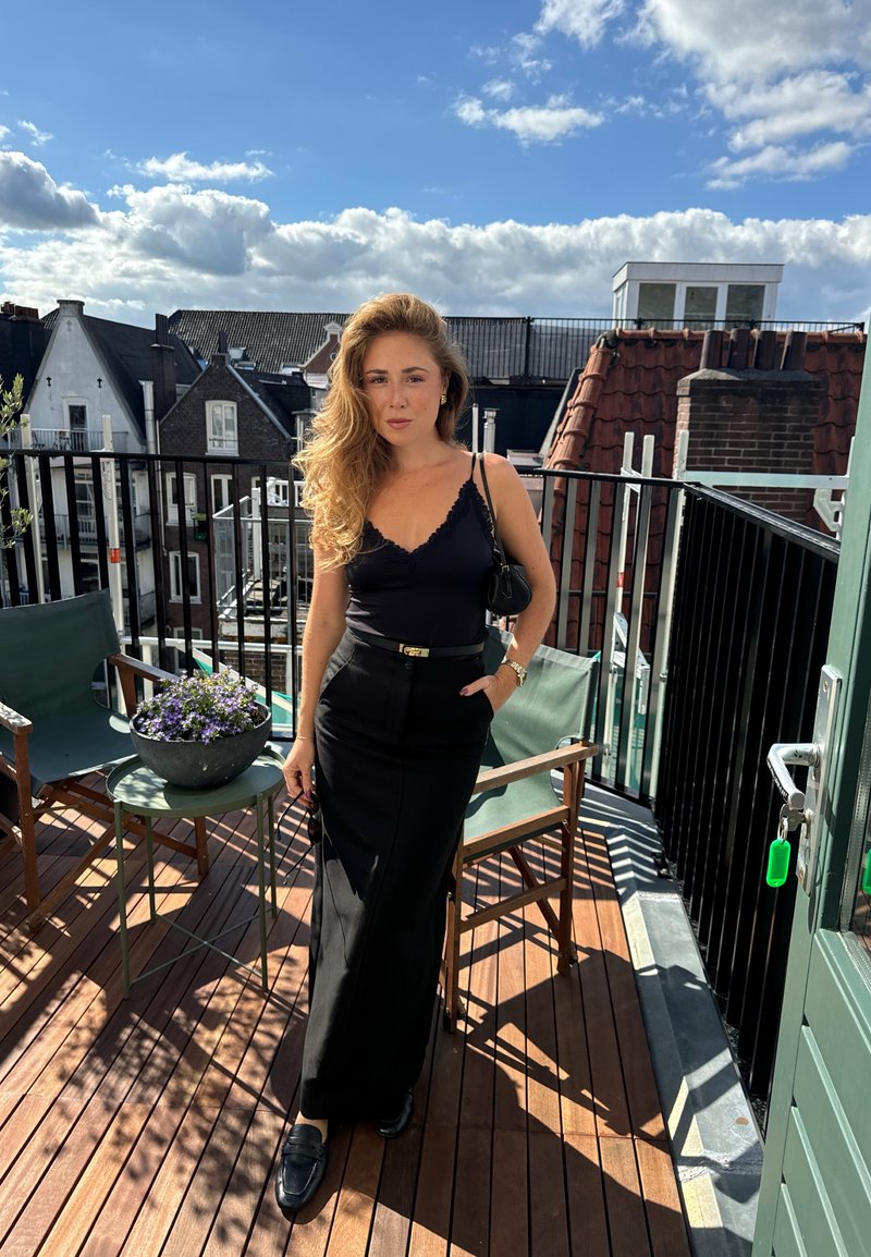 Donna con lunghi capelli ricci, vestita con una maglietta nera e una gonna, si trova su una terrazza sul tetto con pavimento in legno, sedie e fiori in vaso sotto un cielo nuvoloso.