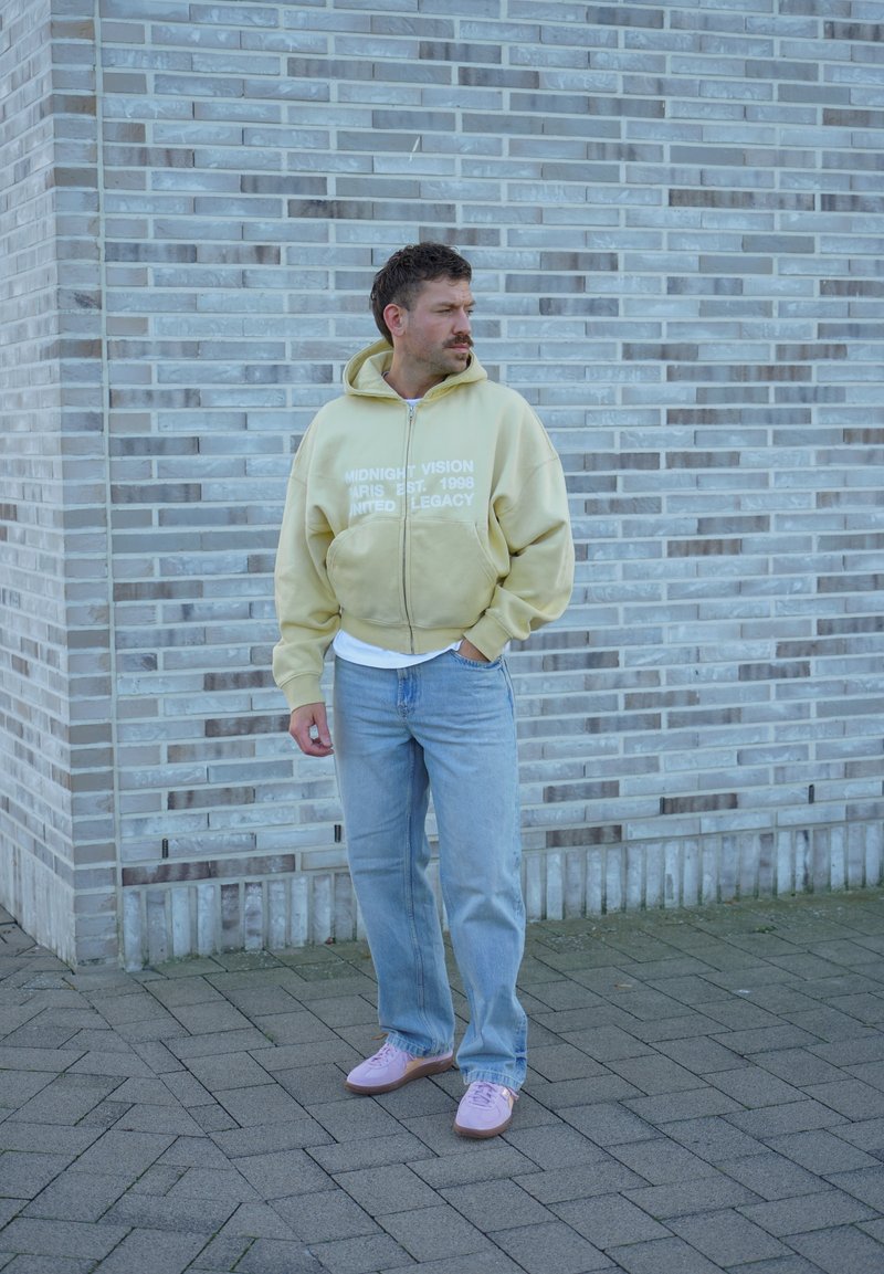 Un homme portant un sweat à capuche jaune pâle zippé, un jean bleu et des baskets roses se tient sur un trottoir pavé et regarde sur le côté, devant un mur de briques gris.