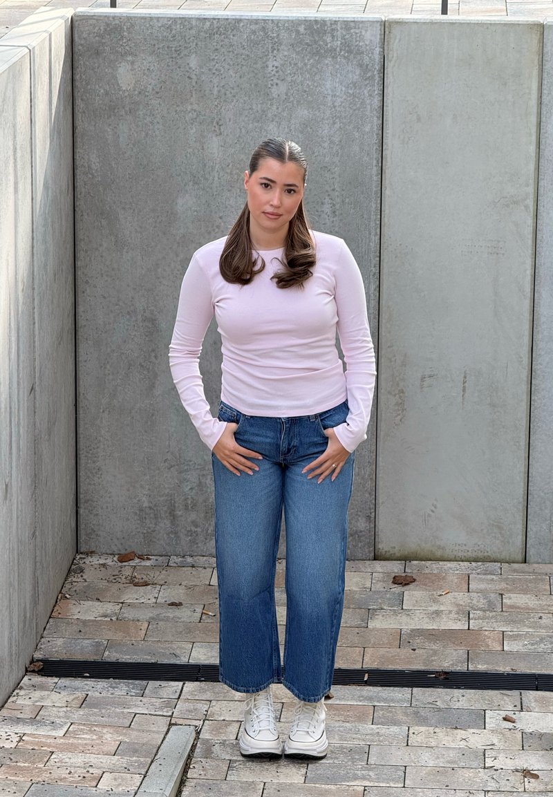 Jeune femme aux longs cheveux bruns portant une chemise rose clair à manches longues, un jean bleu et des baskets blanches, debout contre un mur de béton gris.