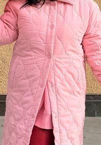 Personne portant un long manteau matelassé rose à boutons-pression par-dessus un vêtement rose plus clair et un pantalon rouge foncé.