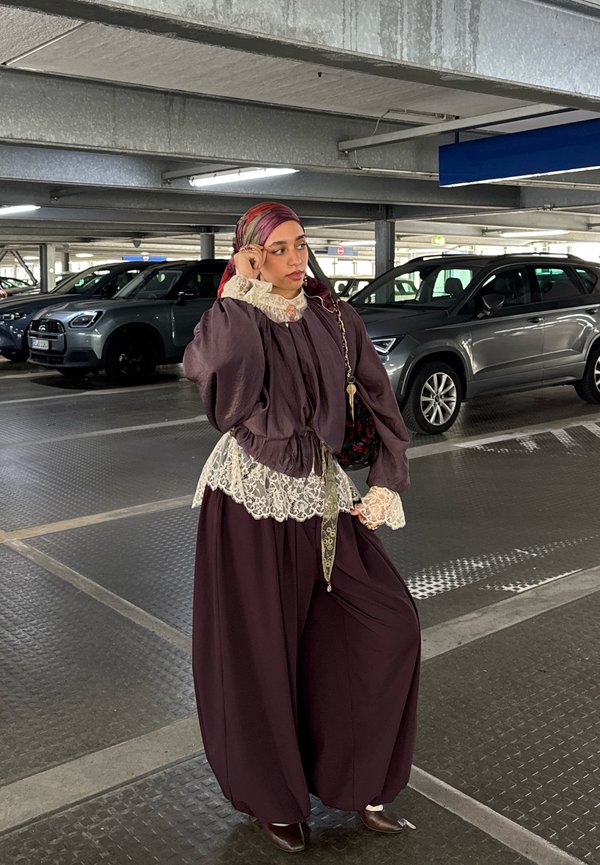 Žena v fialovom oblečení s čipkovými detailmi a farebnou šatkou na hlave stojí v viackomorovej parkovacej garáži blízko zaparkovaných áut.
