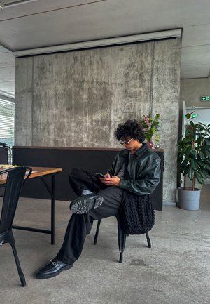 Mocasines de cuero negro, pantalones negros y una chaqueta de cuero verde oscuro. Persona sentada en una silla negra en un interior moderno y minimalista.