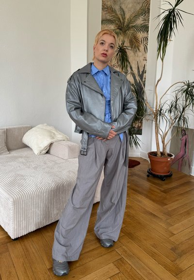 Chaqueta de moto de cuero sintético plateado, camisa azul clara, pantalones de pierna ancha gris y zapatos grises. Entorno interior con plantas y un sofá.