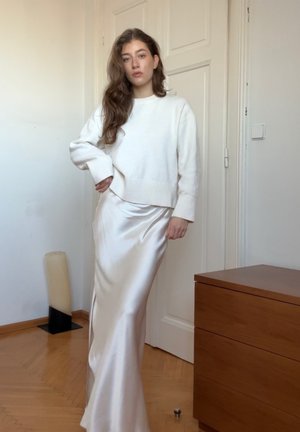 Suéter de punto blanco y falda satinada color crema con abertura lateral. Textura suave, corte relajado y silueta moderna, contra un fondo interior claro.