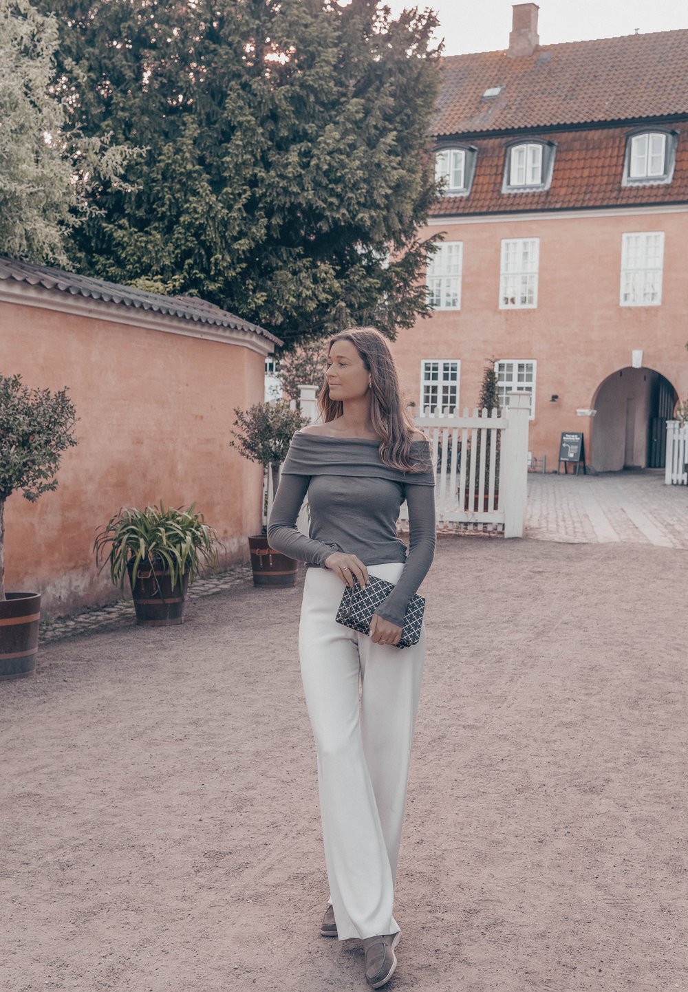 Γκρι τοπ χωρίς ώμους, λευκά παντελόνια με φαρδιά πόδια, κρατάει μια μοτίβο μαύρη τσάντα. Το φόντο περιλαμβάνει γλάστρες με φυτά και ροζ κτίρια.