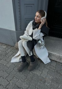 Ανοιχτό γκρι μακρύ παλτό, μαύρο κασκόλ, λευκά παντελόνια, γκρι παντόφλες, ασημένια τσάντα σχήματος καρδιάς. Το μοντέλο κάθεται σε έναν υφασμένο τοίχο.