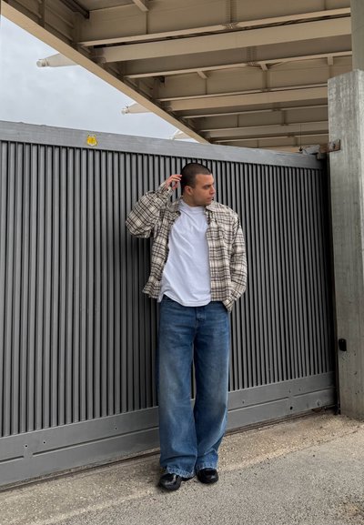Camisa de franela a cuadros sobre una camiseta blanca, combinada con jeans de pierna ancha azul y zapatos negros, posando frente a una puerta gris con listones.