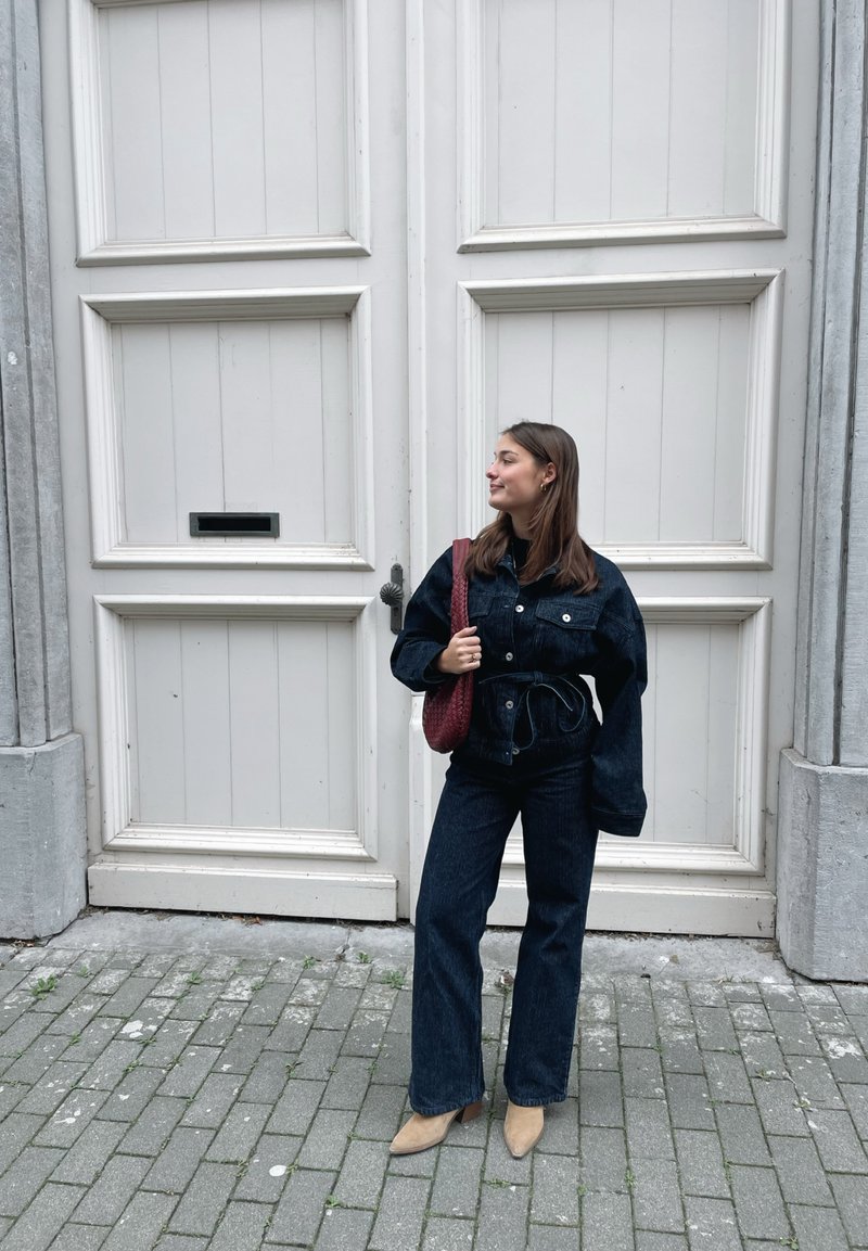 Denim jacket and wide-leg trousers in dark blue denim, complemented by a red woven handbag and beige shoes, set against a pale door.