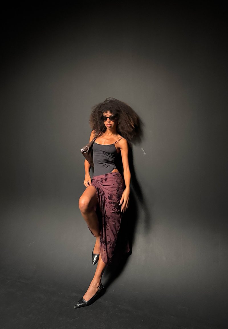 Femme aux cheveux bouclés portant des lunettes de soleil, un haut noir, une jupe violette avec une fente et des chaussures noires, posant devant un fond sombre uni.