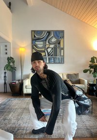 Czarny prążkowany sweter z dzianiny, białe spodnie i czarne buty. Trzyma książki, z widoczną fakturowaną czarną torbą oraz wyposażeniem wnętrza.