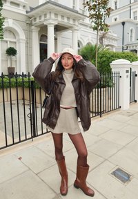 Casaco oversized de pele castanha, gola alta beige com textura canelada, saia de malha a condizer, collants castanhos e botas castanhas até ao tornozelo com um ligeiro salto.