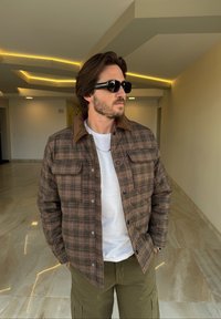 Brun ternet jakke med knaplukning foran og kontrasterende tan krave, båret over en hvid skjorte og kombineret med mørke solbriller.