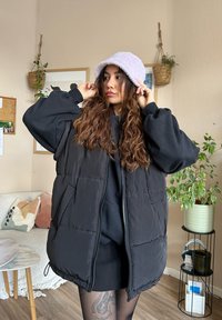 Černá zateplená vesta se zipem a bočními kapsami, navrstvená over černým oblečením. Měkký lila kbelíkový klobouk doplňuje outfit.