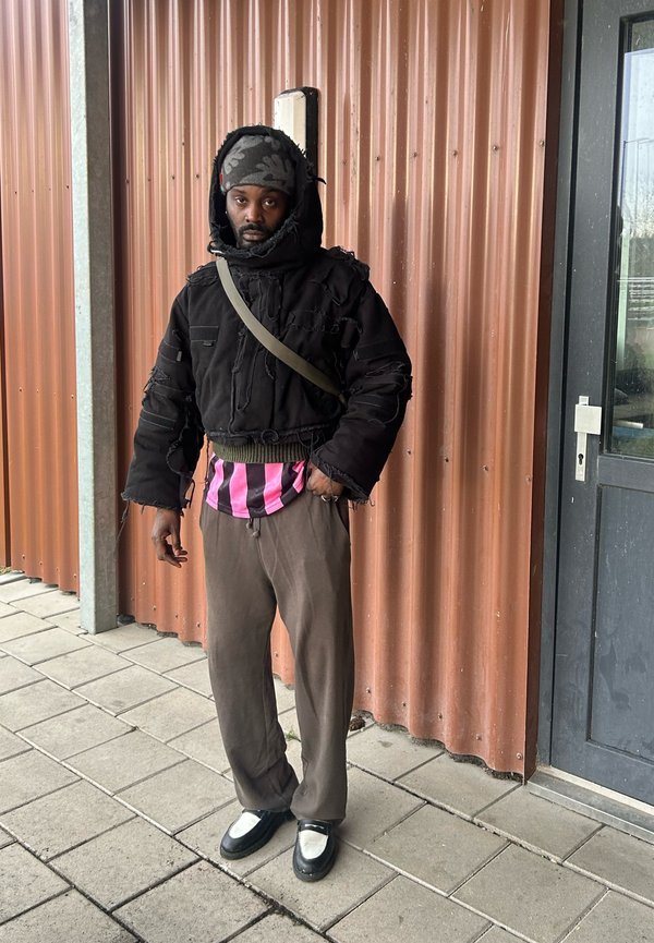 Chaqueta acolchada negra con bordes deshilachados, camisa a rayas rosa y negra debajo, pantalones de pierna ancha marrones y mocasines negros con acentos blancos.