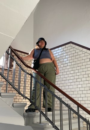Una persona está de pie en una escalera, vistiendo un top corto gris, pantalones oliva y un sombrero tipo cubo negro, con un bolso cruzado.
