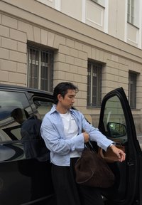 Mladý muž si upravuje rukáv, zatiaľ čo stojí vedľa otvorených dverí auta a v ruke drží veľkú hnedú semišovú tašku na mestkej ulici s béžovou budovou v pozadí.
