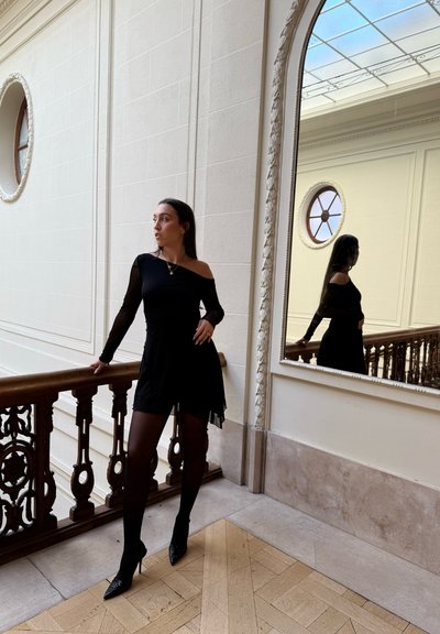 Vestido negro de un solo hombro con mangas largas transparentes, combinado con medias negras y tacones de punta. Barandilla de madera y espejo ornamentado en el fondo.