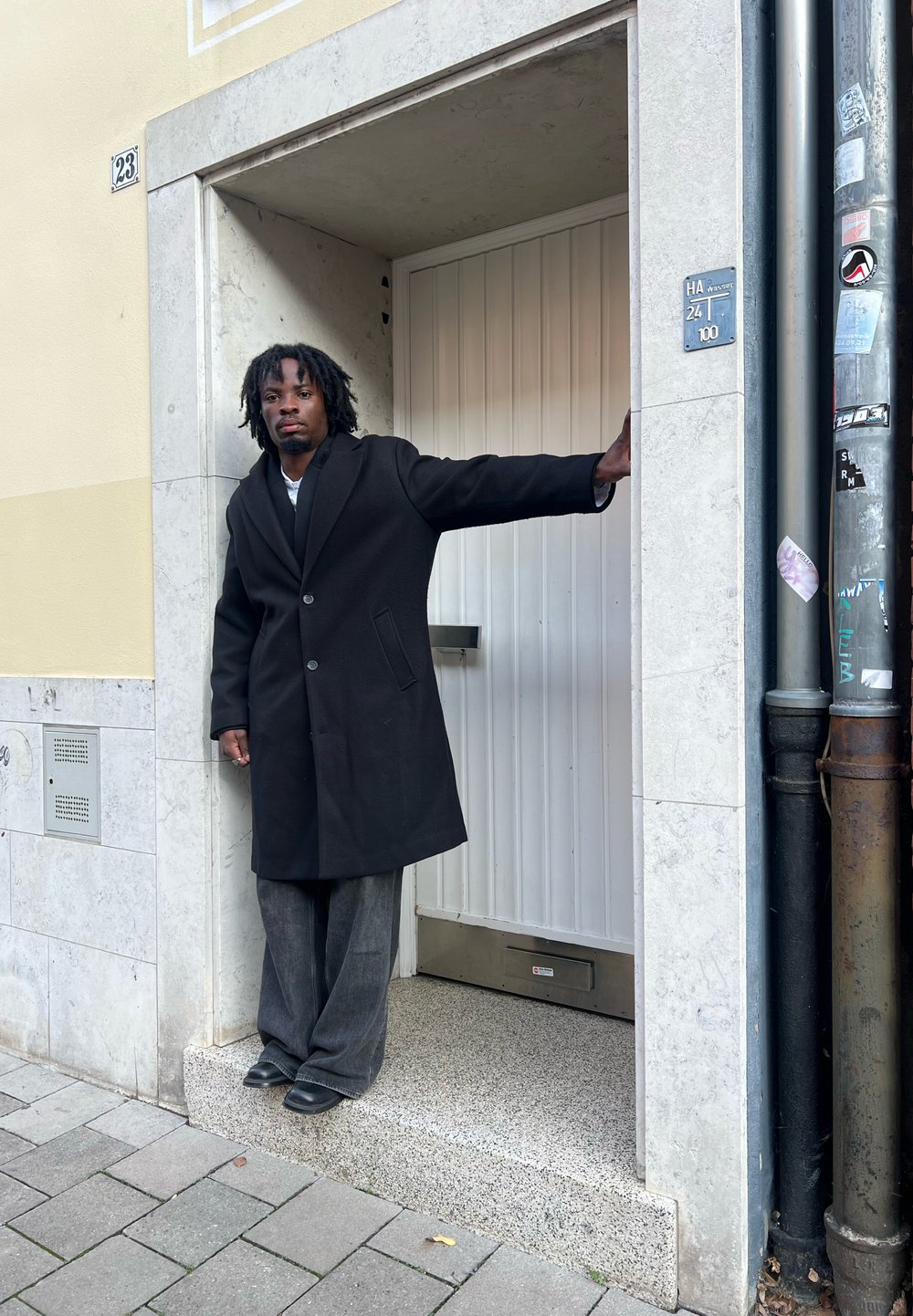 Czarny wełniany płaszcz z wyłogiem, połączony z szerokimi szarymi spodniami. Otoczenie obejmuje wejście z białymi drzwiami i kamienną podstawą.