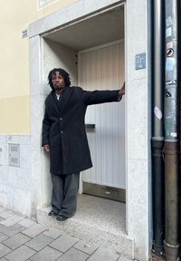 Czarny wełniany płaszcz z wyłogiem, połączony z szerokimi szarymi spodniami. Otoczenie obejmuje wejście z białymi drzwiami i kamienną podstawą.