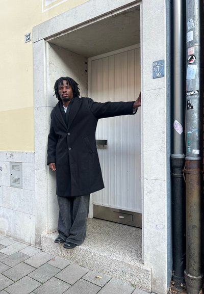 Abrigo negro de lana con cuello con muesca, combinado con pantalones anchos grises. El entorno presenta una puerta blanca con base de piedra.