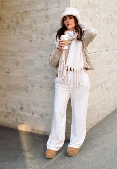 Suéter de punto color blanco roto, bufanda a juego con flecos, pantalones blancos de pierna ancha y botas marrones. Sosteniendo un taza de café contra una pared de piedra texturizada.