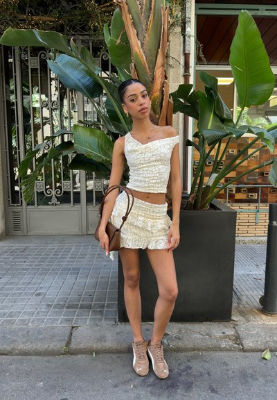 Conjunto blanco texturizado de dos piezas con un top corto y una falda mini. Un bolso marrón y zapatillas beige completan el look. Plantas verdes en el fondo.