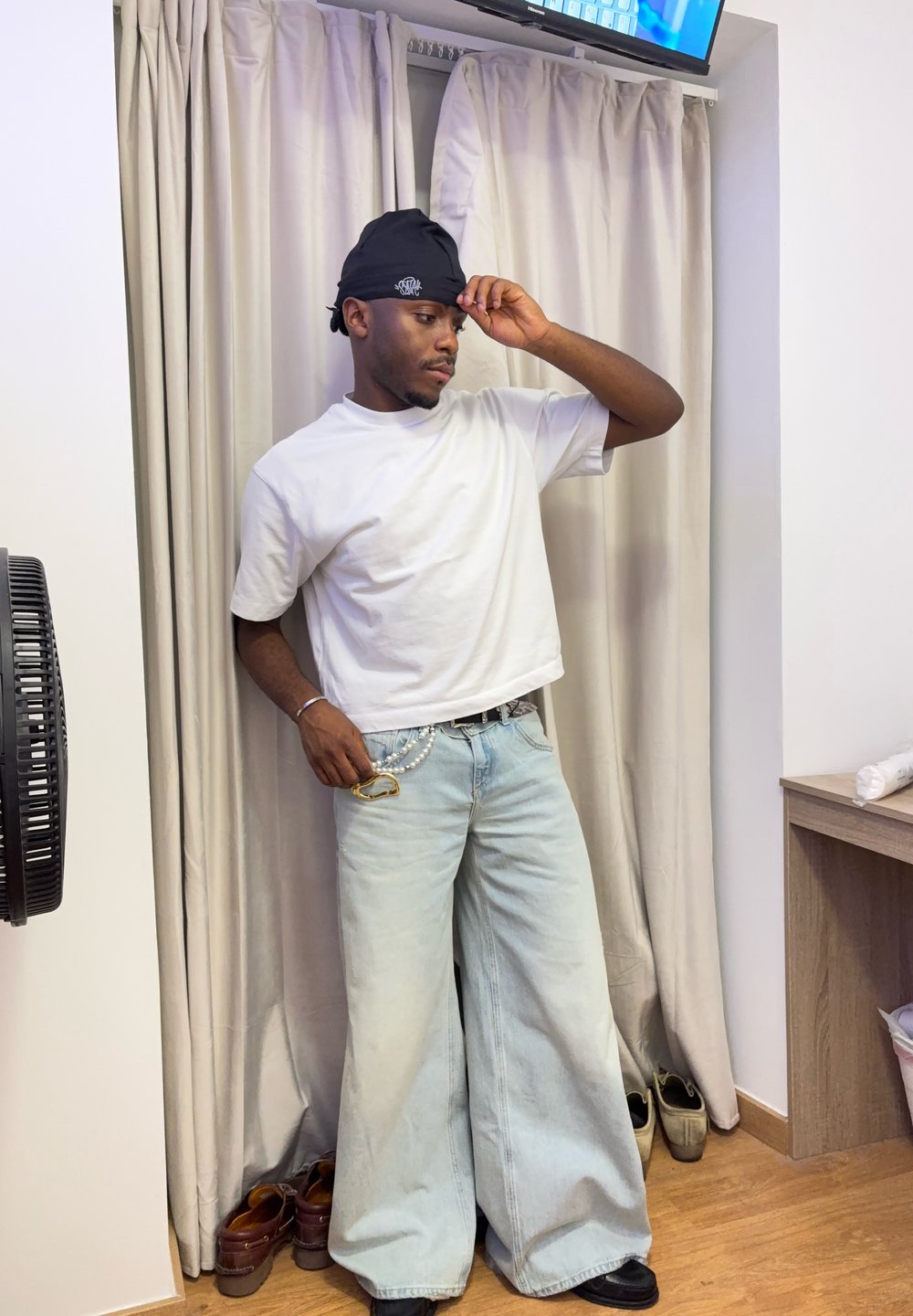 Witte t-shirt met korte mouwen, lichtblauwe wijde jeans, versierd met een zilveren ketting, zwarte pet en zwarte schoenen. Neutrale achtergrond en een ventilator.