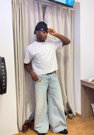 Camiseta blanca de manga corta, jeans de pierna ancha azul claro, adornados con una cadena plateada, gorra negra y zapatos negros. Fondo neutro y ventilador.