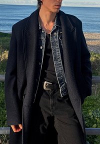 Manteau en laine noire porté sur une veste en denim, chemise côtelée noire et pantalon noir. Boucle de ceinture argentée mettant en valeur la taille. Arrière-plan côtier visible.