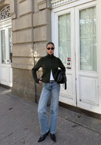 Mulher com um suéter com fecho éclair verde escuro, jeans azuis, botas pretas de bico fino e óculos de sol, segura uma bolsa preta enquanto está em uma calçada da cidade.