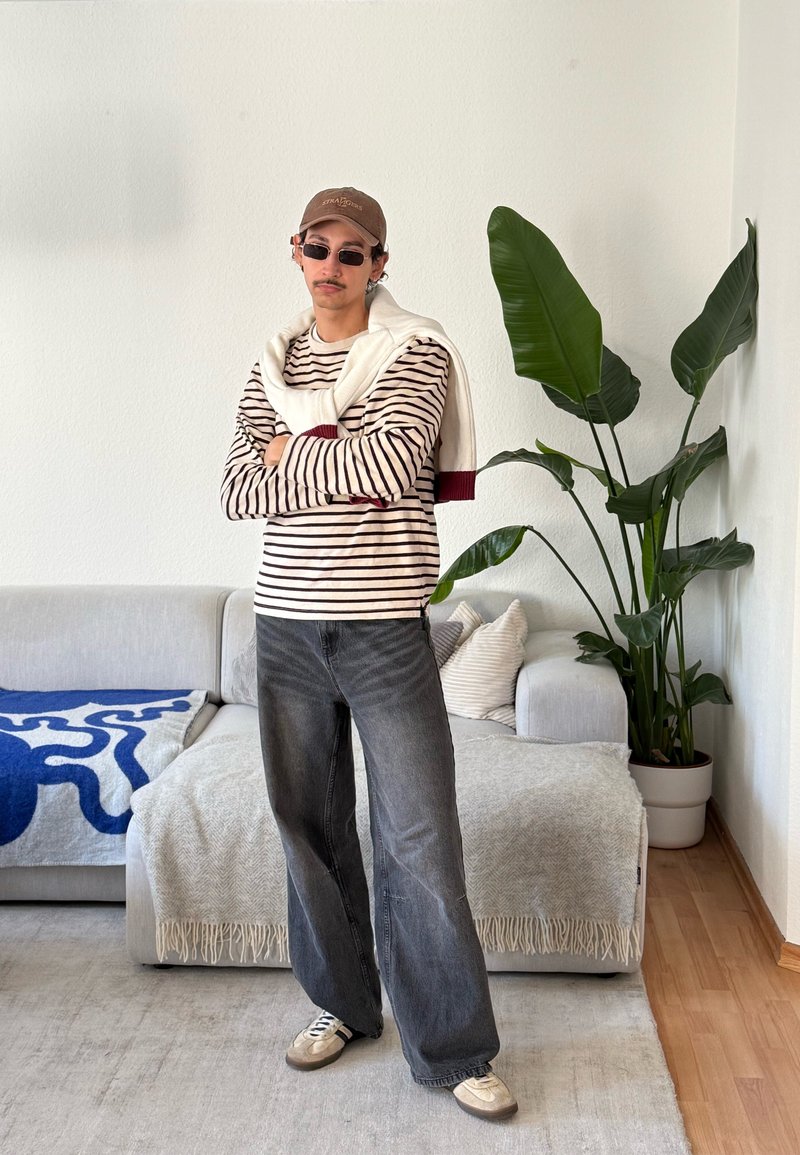 Homme portant une chemise rayée, un jean sombre à jambes larges, une casquette et des lunettes de soleil, debout les bras croisés dans un salon avec un canapé et une grande plante verte.
