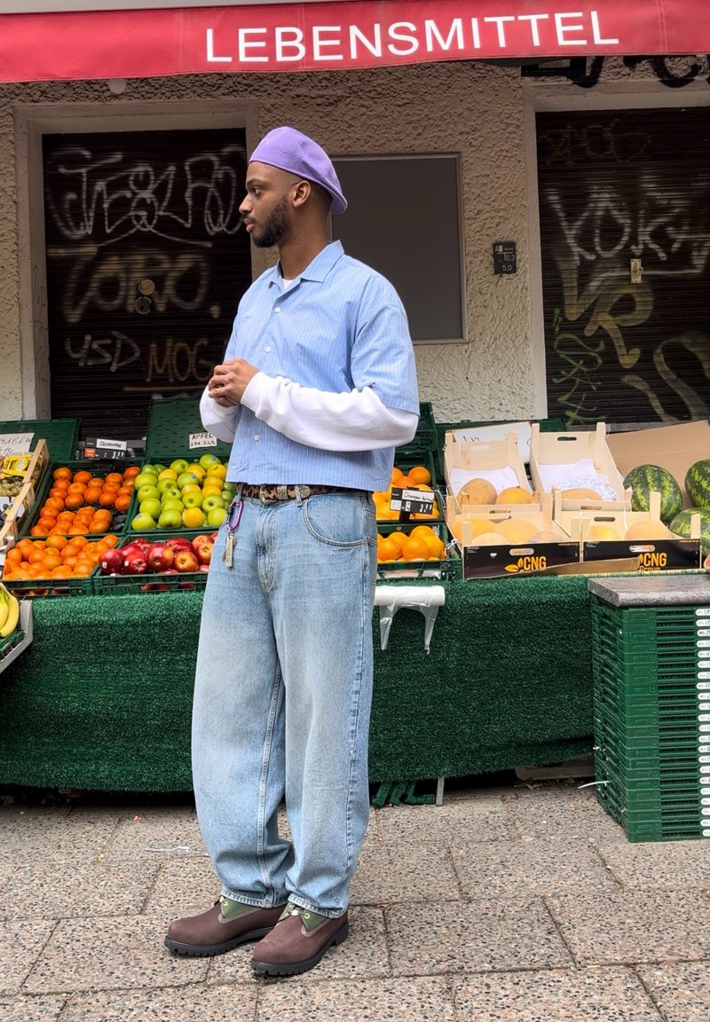 Μπλε και λευκή ριγέ κοντομάνικη μπλούζα, τοποθετημένη πάνω από μια λευκή μακρυμάνικη μπλούζα. Απαλά μπλε παντελόνια με φαρδιά γραμμή, καφέ παπούτσια με πράσινες λεπτομέρειες.