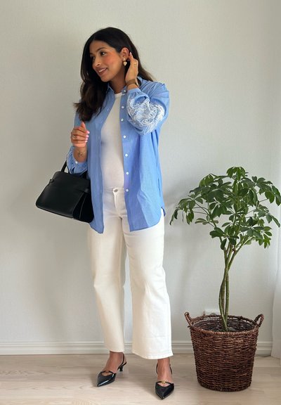 Camisa de botones azul claro con bordados florales, camiseta ajustada blanca, pantalones de pierna ancha blancos, bolso negro y tacones negros de punta.