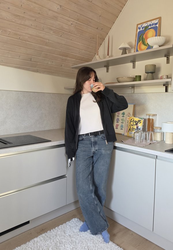 Young woman in casual clothes drinking from a small cup in a modern kitchen with light wood ceiling and white cabinets.